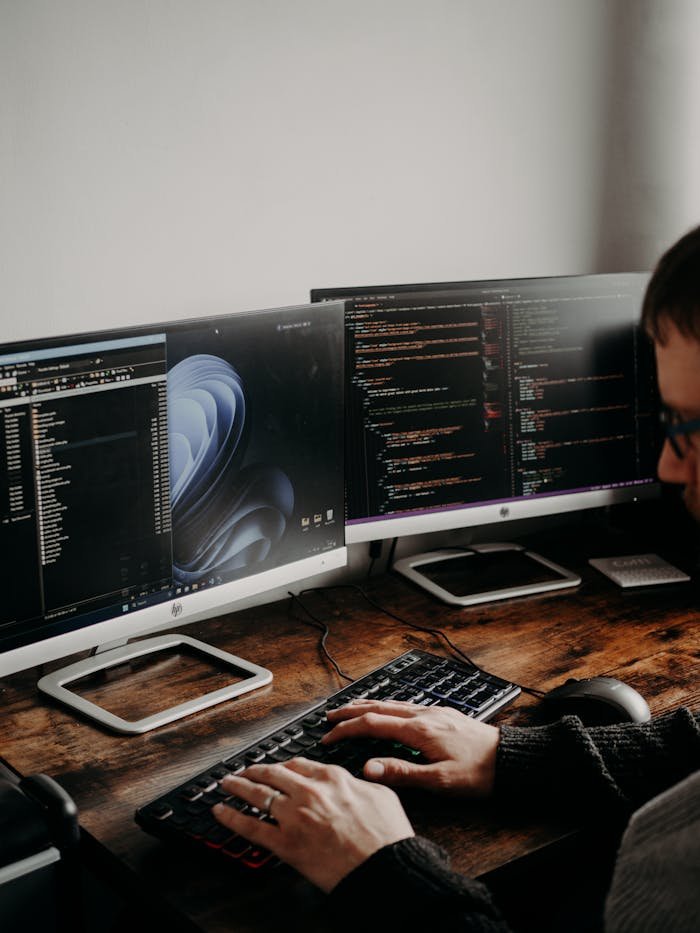 A man working on website design and coding at a home office with a dual monitor setup.